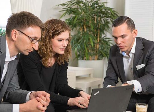 En man och en kvinna sitter vid ett lägre bord på ett kontor och ser ner på en laptop samtidigt som en tredje person sitter och förklarar något som syns på skärmen.