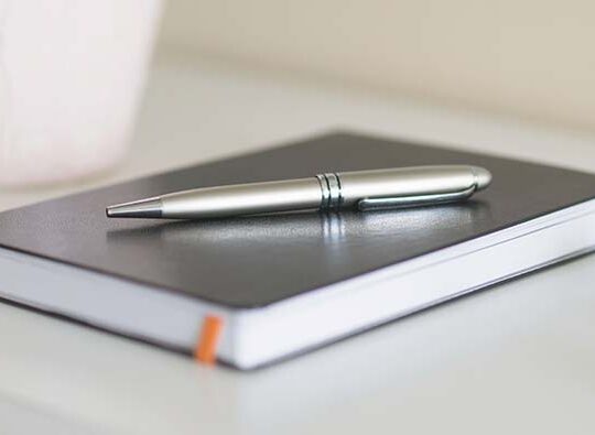 Närbild på ett svart anteckningsblock som ligger på ett bord med en kulspetspenna i metall på.