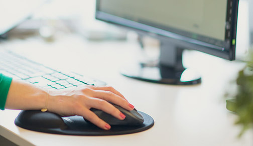 En kvinnlig hand med rödmålade naglar håller i en datormus vid ett tangentbord på ett vitt skrivbord. Framför står en datorskärm.