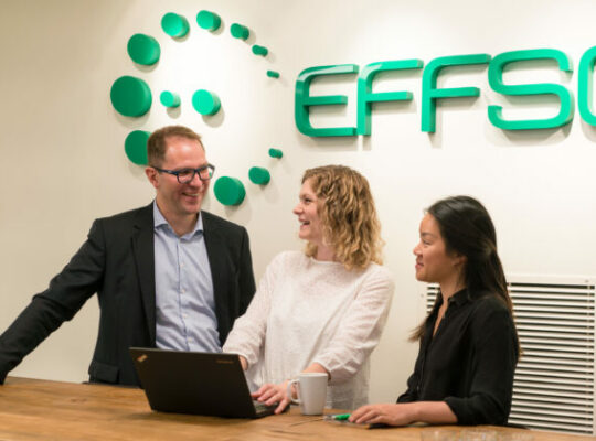 Tre personer, en man och två kvinnor står vid ett bord, framför en laptop och pratar och ser glada ut. Bakom dem på väggen står det EFFSO i stora gröna bokstäver.