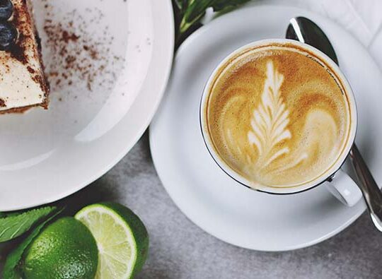 Bild uppifrån på en tjusig frukost med en kopp cappuccino, ett fat med något sött på och grönablad och frukter.