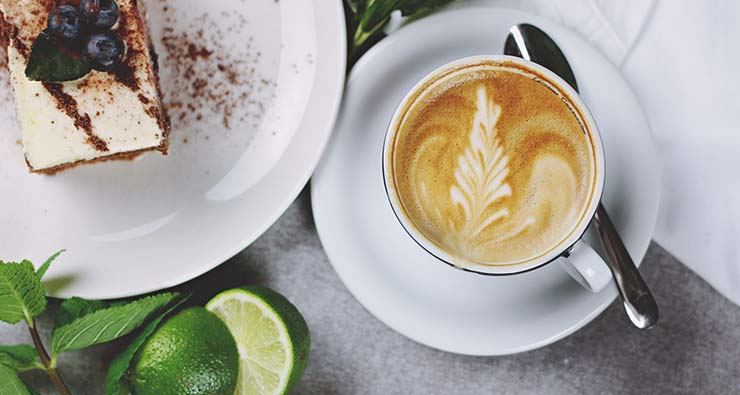 Bild uppifrån på en tjusig frukost med en kopp cappuccino, ett fat med något sött på och grönablad och frukter.