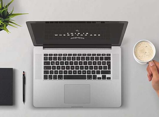 Bild uppifrån på ett välstädat skrivbord dä man ser en laptop, ett anteckningsblock, en penna, en liten grön krukväxt och en hand som håller en kaffekopp.