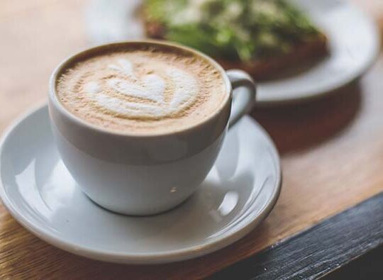 Närbild på frukost med en kopp cappuccino och ett fat med något grönt på.