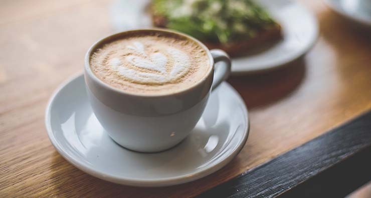 Närbild på frukost med en kopp cappuccino och ett fat med något grönt på.