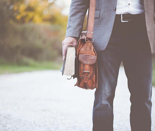 Nederdelen av en man i kavaj och mörka byxor som står utomhus och håller i en tjock bok. På axeln hänger en brun väska.