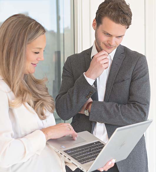 En kvinna i långt hår och ljus skjorta håller i en laptop och visar något för en manlig kollega i grå kavaj.