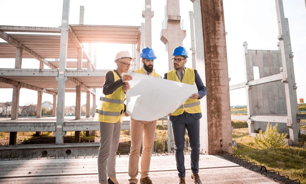Tre personer iförda hjälmar står på en byggarbetsplats och tittar tillsammans på en ritning.