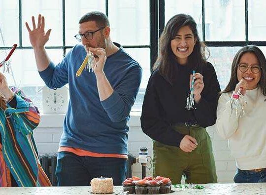Fyra personer står på rad framför kameran och firar något med ballonger och tårta.