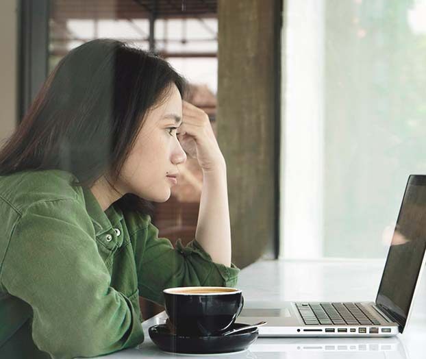 Ung kvinna med brunt hår är fotad från sidan när hon läsaer på en bärbar dator, Framför sig har hon en kaffe- eller tekopp.