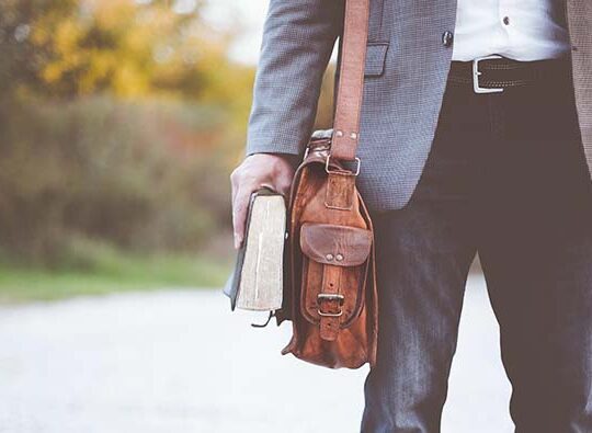 Detaljbild där man ser en man till höger som är på väg någonstans med en axelremsväska och en bok i handen.