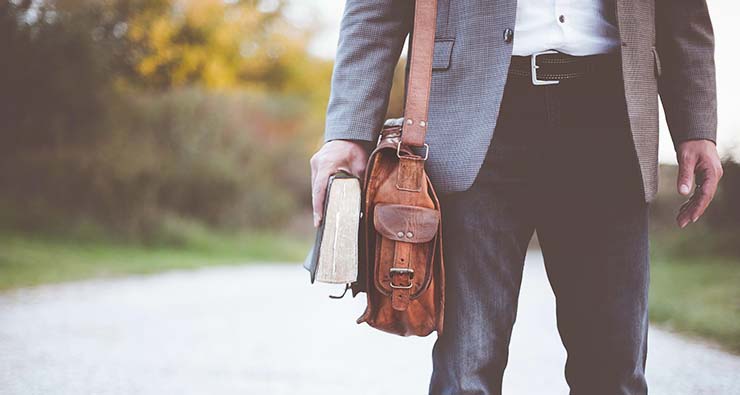 Detaljbild där man ser en man till höger som är på väg någonstans med en axelremsväska och en bok i handen.