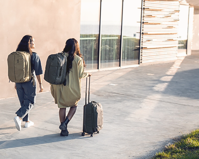 Två kvinnor promenerar iväg med resebagage i form av ryggsäckar och en kabinväska.