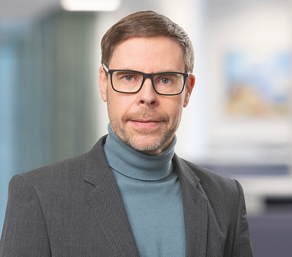 Inomhusporträtt på en man i glasögon, blå polotröja och mörkgrå kavaj.