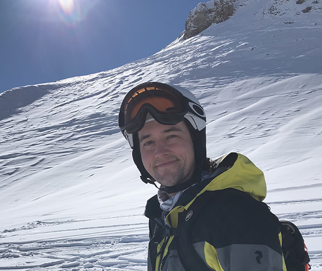 Foto på en man i skidhjälm som står i en nerförsbacke och med sol och blå himmel bakom sig.