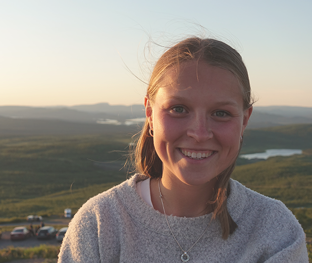 En ung kvinna står i ett landskap och ler mot kameran. Bakom henne ser man vacker natur och solen lyser upp hennes ansikte.