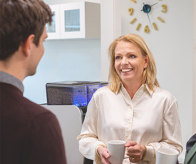 En blond kvinna i vit blus står vid en kaffemaskin och pratar med en yngre kollega.