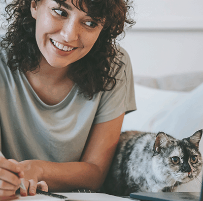 Mörkhårig ung kvinna ser snett ner mot en datorskärm samtidigt som hon skriver med en penna på ett papper bredvid. På bordet sitter även en katt och tittar på samma skärm.