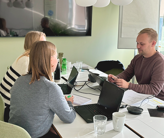 Två yngre kvinnor och en man sitter och arbetar vid ett bord i ett grönt konferensrum.