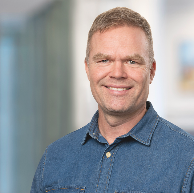 Porträtt på en man i jeansskjorta som ser in i kameran och ler.