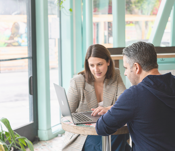 En man och en kvinna sitter till höger i bild vid ett cafébord och ser ner på en laptop.