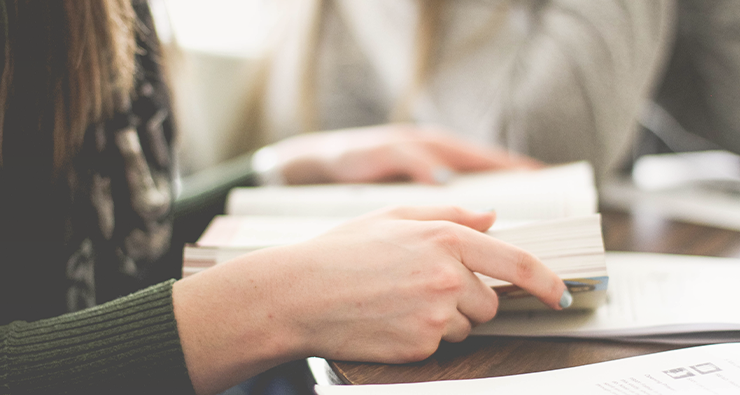 Närbild på två kvinnohänder som bläddrar i en bok. Kvinnan, i profil, sitter vid ett bord.