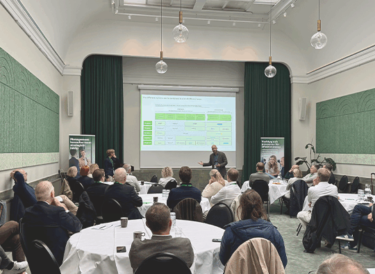 En samling personer sitter vid runda bord på en konferens och lyssnar på två talare, bild tagen bakifrån.