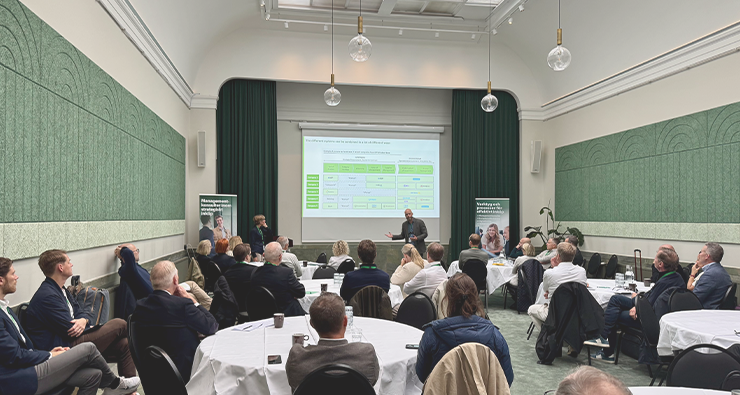 En samling personer sitter vid runda bord på en konferens och lyssnar på två talare, bild tagen bakifrån.