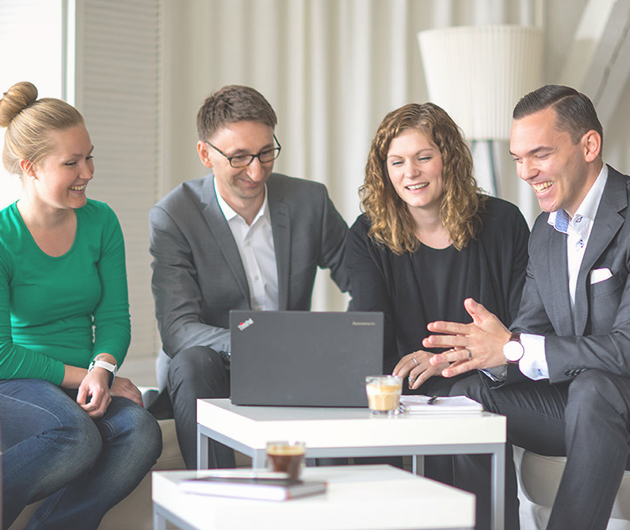 Två män och två kvinnor sitter ner och tittar mot en och samma bärbar dator som står på ett soffbord. De har kul ihop.