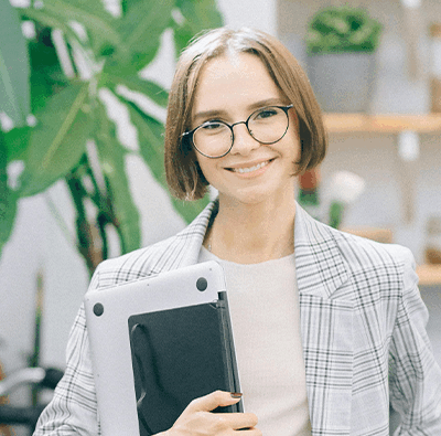 Ung kvinna i page, kavaj och glasögon håller i en dator och ler in i kameran. Bakom grönskande växt och hyllor.