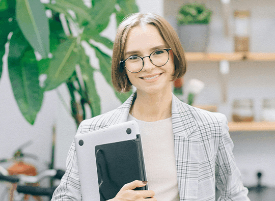 Ung kvinna i page, kavaj och glasögon håller i en dator och ler in i kameran. Bakom grönskande växt och hyllor.