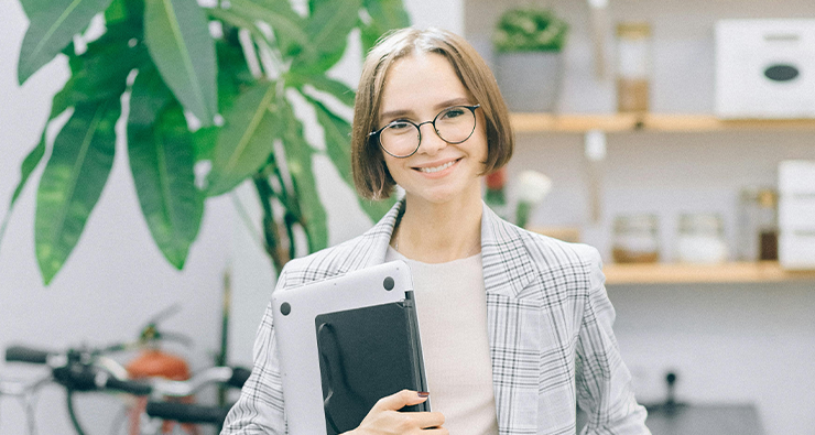 Ung kvinna i page, kavaj och glasögon håller i en dator och ler in i kameran. Bakom grönskande växt och hyllor.