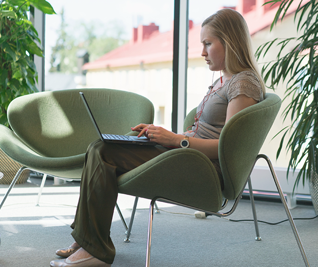 Ung kvinna syns från sidan sitta koncentrerat och titta på sin laptop i en grön loungefåtölj. Gröna växter och fönsterparti i bakgrunden.