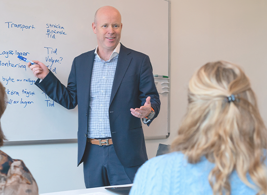 En man i blå skjorta och kostym står och pekar mot en white boardtavla och pratar inför en sittande grupp människor.