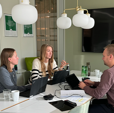 Två kvinnor och en man sitter inne i ett grönt konferensrum och jobbar runt ett bord.