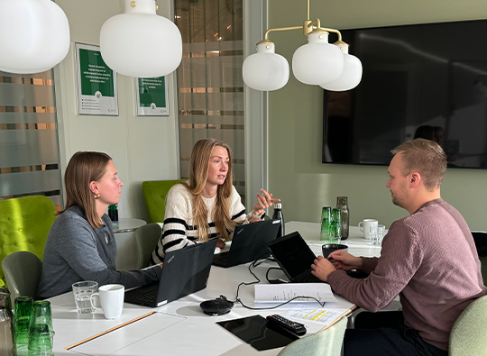 Två kvinnor och en man sitter inne i ett grönt konferensrum och jobbar runt ett bord.