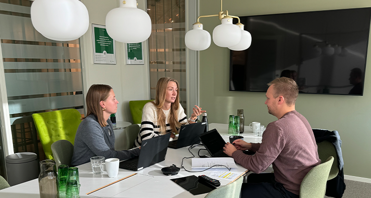 Två kvinnor och en man sitter inne i ett grönt konferensrum och jobbar runt ett bord.