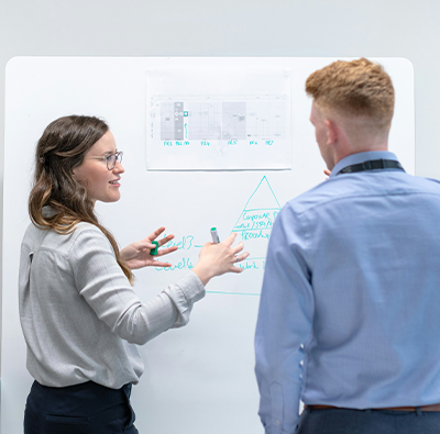 En man och en kvinna ses bakifrån stå vid en white boardtavla med grafer och diskuterar något.