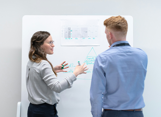 En man och en kvinna ses bakifrån stå vid en white boardtavla med grafer och diskuterar något.