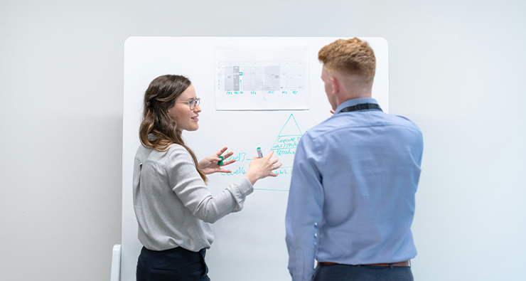 En man och en kvinna ses bakifrån stå vid en white boardtavla med grafer och diskuterar något.