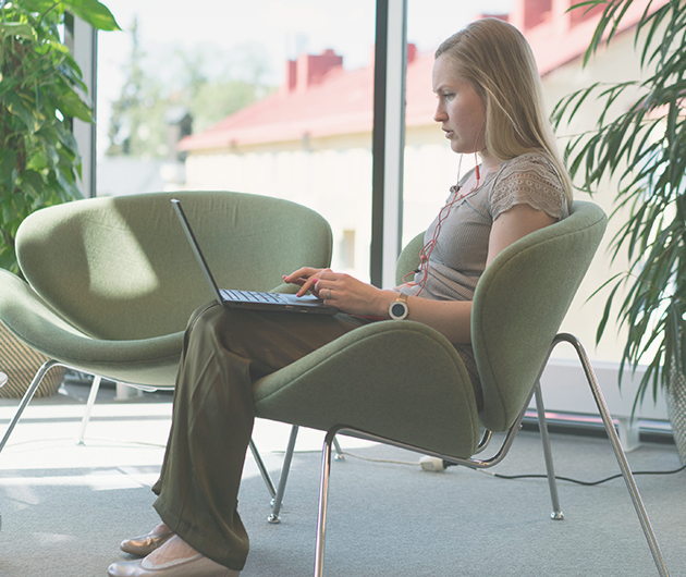Ung kvinna syns från sidan sitta koncentrerat och titta på sin laptop i en grön loungefåtölj. Gröna växter och fönsterparti i bakgrunden.