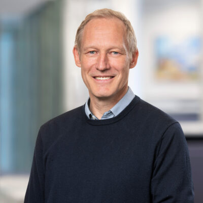 Porträtt på ljushårig man med ljsublå skjorta och marinblå tröja som ser rakt in i kameran och ler.