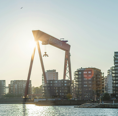 Bild på kranar och flerfamiljshus i Göteborgs hamn, fotat i motljus en sommarkväll.