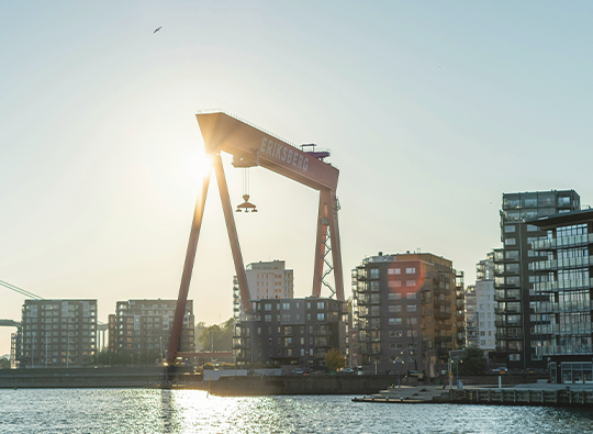 Bild på kranar och flerfamiljshus i Göteborgs hamn, fotat i motljus en sommarkväll.