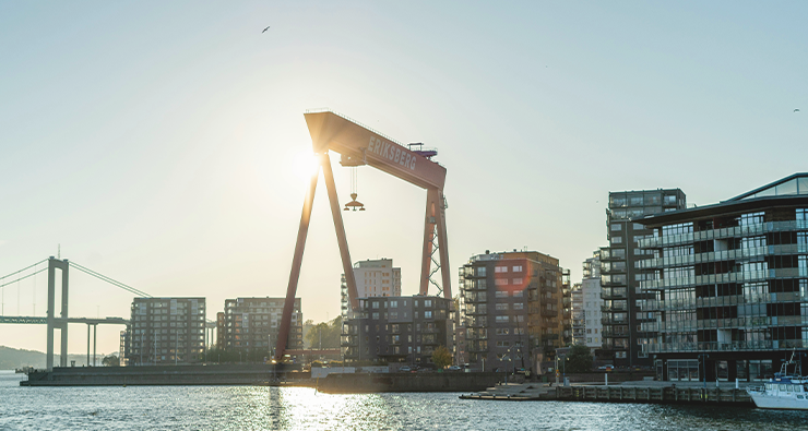 Bild på kranar och flerfamiljshus i Göteborgs hamn, fotat i motljus en sommarkväll.