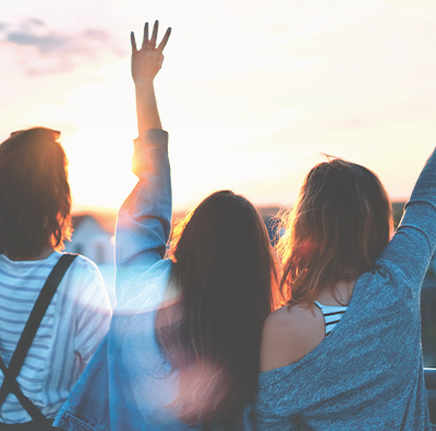 Tre yngre kvinnor syns bakifrån vifta och vinka upp mot en vacker solnedgångshimmel.