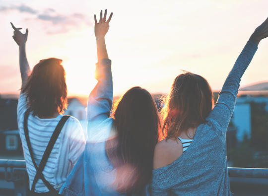 Tre yngre kvinnor syns bakifrån vifta och vinka upp mot en vacker solnedgångshimmel.