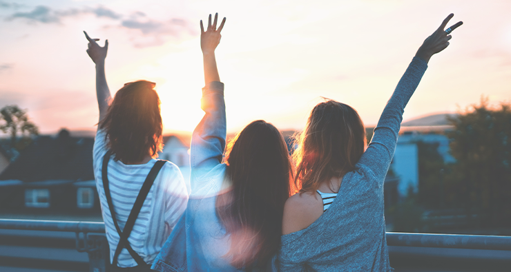 Tre yngre kvinnor syns bakifrån vifta och vinka upp mot en vacker solnedgångshimmel.