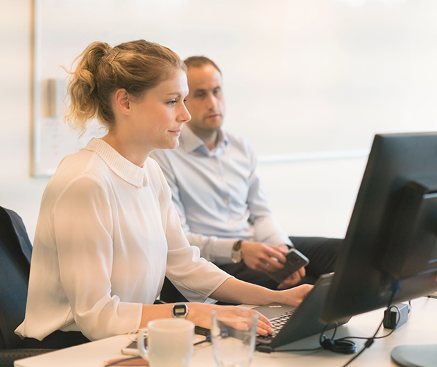 En kvinna i vit blus i förgrunden sitter framför en datorskärm, i i bakgrunden sitter en man och tittar på samma skärm.