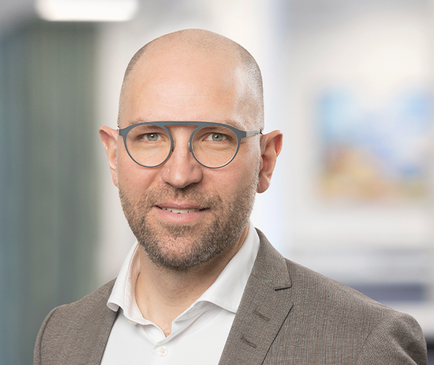 Porträtt inomhus på en man med glasögon, vit skjorta och brun kavaj.